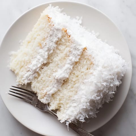 Three-layered homemade Coconut Cake displayed on a white ceramic platter, filled with luscious coconut filling and frosted with billowy meringue.