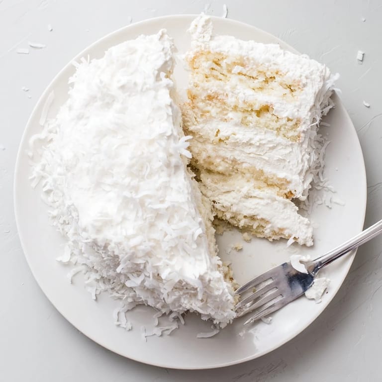 Frosted Coconut Cake slice on a dessert plate showing fluffy white icing and shredded coconut, paired with iced tea for a sweet treat.