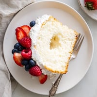 Freshly baked Angel Food Cake with golden crust, served alongside vibrant mixed berries and a fluffy dollop of whipped cream on a dessert plate.