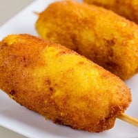 Golden-brown corn dogs on a stick, freshly fried and served with ketchup and mustard.